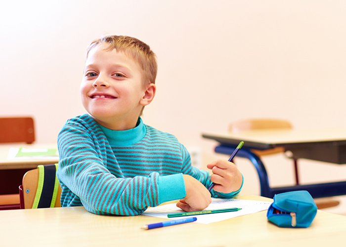 A child smiling whilst holding a pencil and sitting at a desk