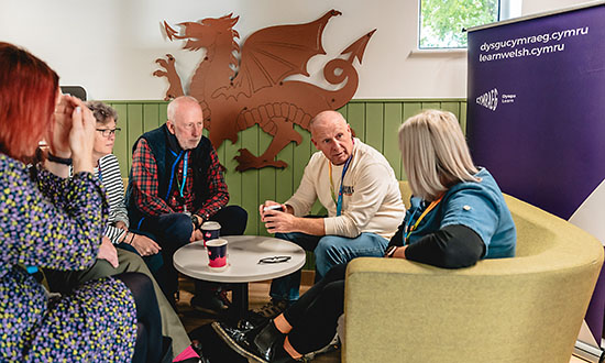 A group of adults sat round a small table with the Welsh flag behind them.