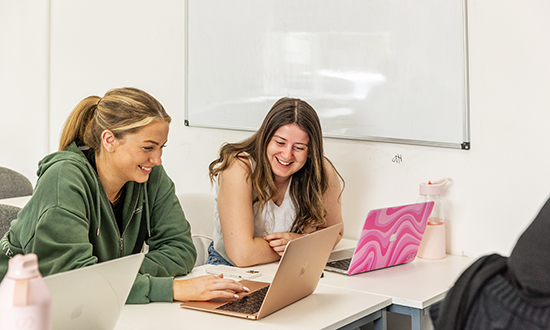 2 young women sat at desks smiling at a laptop screen