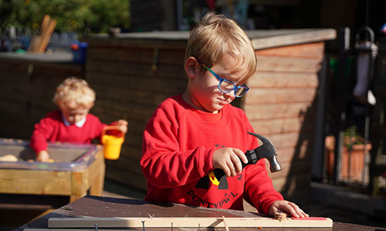 A child in glasses and a red jumper using a hammer on wood