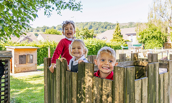 Three primary school students smiling at the camera peaking over a wooden fence.