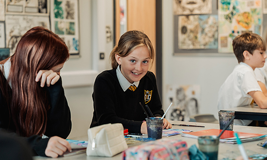 2 young school children in an art class holding paintbrushes, one is looking at the camera