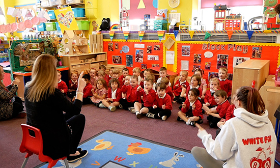 A class of young school pupils sitting on the floor of a classroom with a teacher sitting on a chair