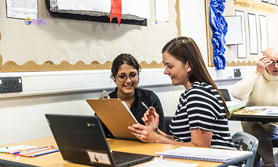 A young woman smiling and talking with another woman looking at a clipboard whilst sitting at a desk
