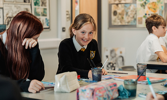 Secondary school children working at desks in a classroom, one looking towards the camera and smiling